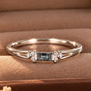 May include: A silver-toned ring featuring a central rectangular baguette-cut dark stone flanked by small clear round stones on each side, presented in a brown velvet-lined box with the text Bareeleganceus printed in gold on the inner lid.