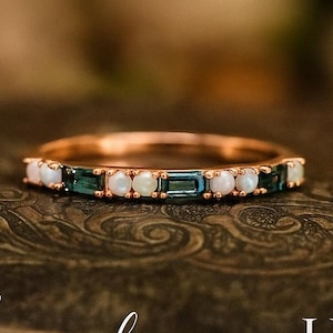Op de afbeelding: Een roségouden ring met afwisselend witte en teal gekleurde edelstenen. De ring ligt op een donkere, getextureerde ondergrond. Een delicaat sieraad.