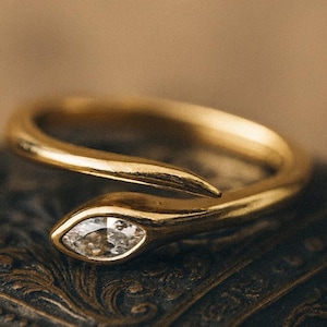 May include: A gold-colored ring in the shape of a snake, with a clear, marquise-cut gemstone as the snake's eye. The ring is set against a dark, textured surface with ornate detailing.