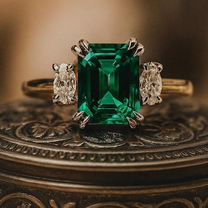 Pode incluir: Um anel de ouro com uma grande gema verde esmeralda, ladeada por dois diamantes ovais menores. O anel está sobre uma caixa de metal decorativa de estilo antigo. O texto "Bare Eleganceus" é visível.