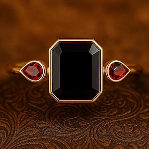 May include: A gold ring featuring a large, rectangular black gemstone as the centerpiece, flanked by two smaller, teardrop-shaped red gemstones. The ring is set against a textured, brown background. The text "baree eleganceus" is visible.