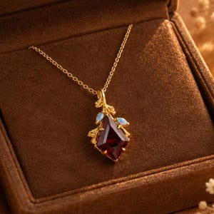 May include: A gold necklace featuring a kite-shaped dark red gemstone pendant with small blue accents, presented in a brown velvet box with the text Bareeleganceus printed in gold on the inner lid, showcasing a delicate chain and branch-like setting.