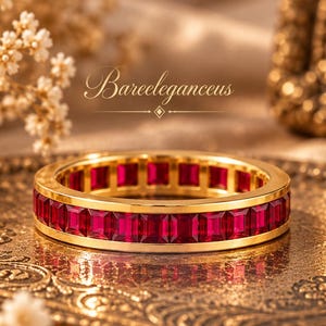 May include: A gold eternity band featuring a continuous row of vibrant red, rectangular-cut gemstones set within a polished metal frame. The ring is centred below the text Bareleganceus, showcasing a sleek, uniform design with deep crimson stones.