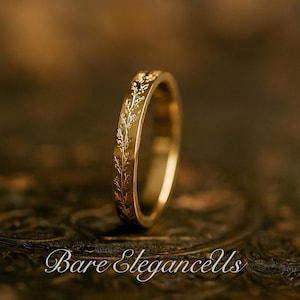 Könnte beinhalten: Ein goldfarbener Ring mit einem floralen Muster, das um das Band geätzt ist. Der Ring wird auf einer dunklen, strukturierten Oberfläche präsentiert. Der Schriftzug "Bare Elegancells" ist unten im Bild zu sehen.