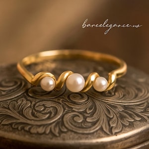 May include: A gold ring featuring three pearls. The ring has a distinctive, wavy design, with the band holding the pearls. The pearls are white and round. The ring is displayed on a decorative, silver-coloured surface. The text "bareelegance.us" is in the upper right corner.