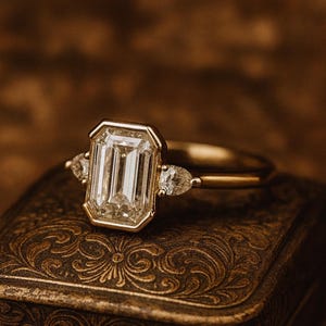 May include: A gold ring featuring a large, rectangular, clear gemstone, with two smaller pear-cut stones either side. The ring sits on a detailed, antique-style box with a brown, textured surface.