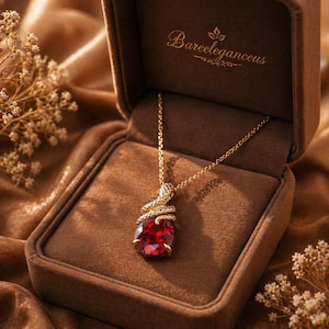 May include: A gold-colored necklace with a large, oval, red gemstone pendant, presented in a brown jewelry box. The pendant is encircled by a snake design. The box is lined with brown velvet and has the text "Bareeleganceus" on the inside.