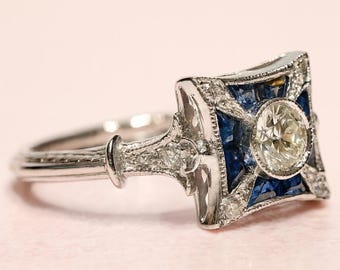 May include: A silver ring with a square, art deco-style setting. The setting features a central, round, clear gemstone surrounded by blue gemstones and smaller clear stones. The ring is on a light pink background.