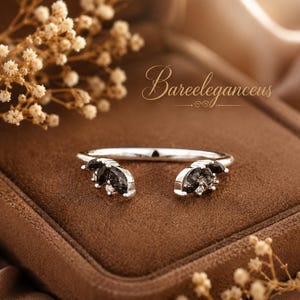 Puede incluir: Un anillo de plata con piedras negras y pequeños acentos blancos. El anillo tiene un diseño abierto con las piedras preciosas agrupadas en cada extremo. El anillo se exhibe sobre una superficie marrón con flores secas.
