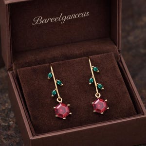 Puede incluir: Un par de pendientes colgantes dorados con diseño floral, presentados en una caja de joyería marrón. Cada pendiente presenta una gema roja hexagonal en la parte inferior, con detalles verdes en forma de hoja y el texto "Bareelganceus" en la caja.