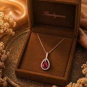 Puede incluir: Un collar dorado con un colgante de gema roja en forma de lágrima, rodeado de pequeñas piedras blancas, descansa en una caja de terciopelo marrón abierta. El texto "Bareeleganceus" es visible en la caja.