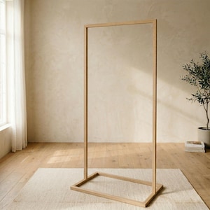 Puede incluir: Un perchero rectangular de madera minimalista. El marco es de madera de color claro y se apoya sobre una base rectangular. El perchero está vacío, y el fondo presenta una pared de tonos neutros, una alfombra y una planta en maceta.