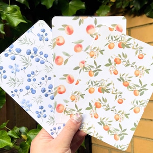 May include: Three square cards with botanical designs. One card features blue berries and green leaves on a white background. The other two cards display peaches and orange blossoms on a white background. The cards are held in a hand.