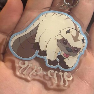 Könnte beinhalten: Ein klarer Acryl-Schlüsselanhänger mit einem Cartoon-Charakter. Der Charakter hat weißes Fell, braune Details und eine blaue Umrandung. Er enthält einen silbernen Verschluss und ein klares Etikett mit stilisiertem Text.