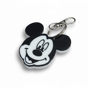 May include: A black, white, and silver Mickey Mouse keychain. The keychain features a smiling Mickey Mouse face with black ears and a white face. The keychain has a silver clasp and ring.