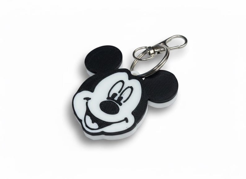 May include: A black, white, and silver Mickey Mouse keychain. The keychain features a smiling Mickey Mouse face with black ears and a white face. The keychain has a silver clasp and ring.