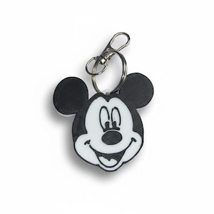 May include: A black and white Mickey Mouse keychain with a silver clasp. The keychain features a smiling Mickey Mouse face with large black ears and a white face. The design is simple and iconic.