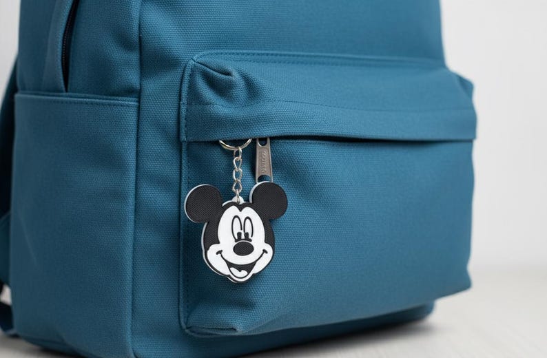 May include: Teal backpack with a front pocket and a Mickey Mouse keychain. The keychain is black and white. The backpack has a zipper on the front pocket.