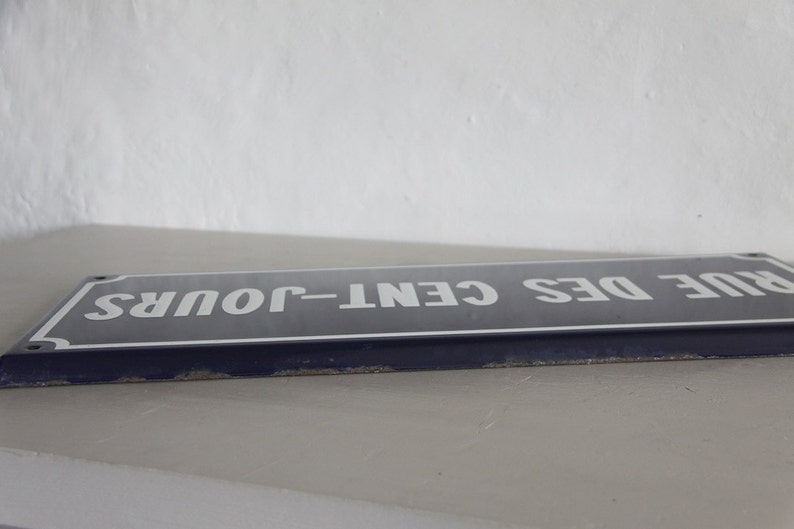 May include: A blue enamel street sign with white lettering that reads "RUE DES CENT-JOURS".