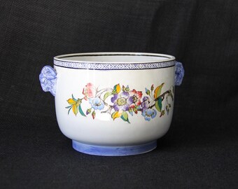 Op de afbeelding: Een witte keramische kom met blauwe en witte rand en bloemmotieven. De kom heeft twee blauwe handvatten in de vorm van leeuwenkoppen.