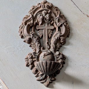 May include: Ornate, brown wall-mounted religious artifact featuring a central cross, surrounded by detailed carvings of leaves, flowers, and cherubic figures. The bottom portion includes a shell-shaped basin. The overall design is symmetrical and detailed.