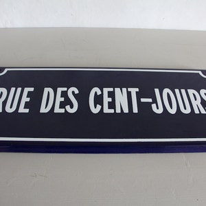 May include: A blue enamel street sign with white lettering that reads "RUE DES CENT-JOURS".