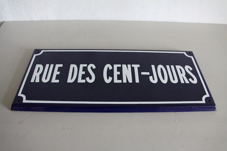 May include: A blue enamel street sign with white lettering that reads "RUE DES CENT-JOURS".