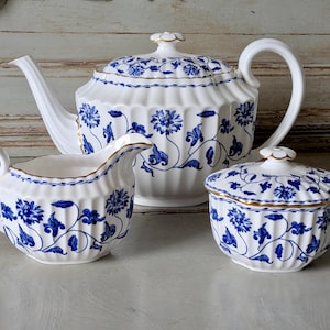 May include: A white porcelain tea set with blue floral patterns. The set includes a teapot, creamer, and covered sugar bowl. The rims are trimmed with gold. The teapot has a curved spout and handle. The creamer has a curved handle and spout.