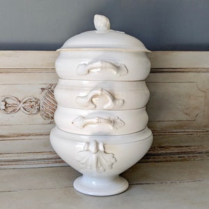 Puede incluir: Un plato de servir de cerámica blanca de varios niveles con detalles ornamentados. El plato está apilado en cuatro niveles y presenta asas decorativas y acentos florales.