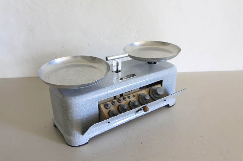 May include: A vintage white balance scale with two silver metal trays and a drawer containing a set of weights. The scale has a wooden handle and a metal lever.