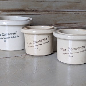 May include: Three white ceramic egg cups with brown lettering. The cups are labeled "La Conserve" and have different sizes: 1/2, 1/4, and 1/4.