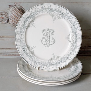 Könnte beinhalten: Vier weiße Keramikteller mit grauem floralem Design am Rand und einem Monogramm in der Mitte. Die Teller sind auf einer hellen Holzoberfläche gestapelt. Jeder Teller hat einen Durchmesser von etwa 25 cm.