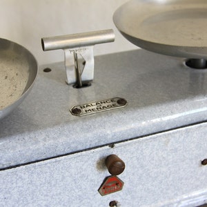 May include: A vintage white enamel kitchen scale with two silver bowls. The scale has a black trim and a label that reads "BALANCE DU MENAGE".