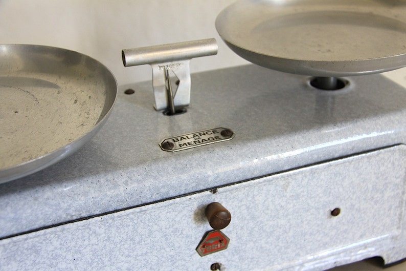 May include: A vintage white enamel kitchen scale with two silver bowls. The scale has a black trim and a label that reads "BALANCE DU MENAGE".