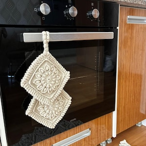 Könnte beinhalten: Zwei quadratische, cremefarbene gehäkelte Topflappen mit einem zentralen Blumenmuster und einem strukturierten Rand. Die Topflappen sind durch eine gehäkelte Schlaufe verbunden und hängen am silbernen Griff eines schwarzen Einbauofens.