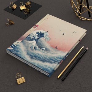 May include: A hardcover journal featuring a print of Hokusai's "The Great Wave off Kanagawa." The cover art depicts a large ocean wave with boats, set against a pink and cream sky. Gold and black pencils and gold binder clips are also visible.