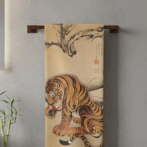 May include: A beige and brown decorative towel with a tiger illustration. The tiger is orange and black with blue eyes. The towel is hanging on a dark brown wooden towel bar in a bathroom setting.