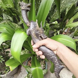 May include: A bronze-colored dagger with dragon-shaped handles and decorative scales. The blade is held in a hand, with lush green foliage in the background. The dagger's design features intricate details and a curved blade.