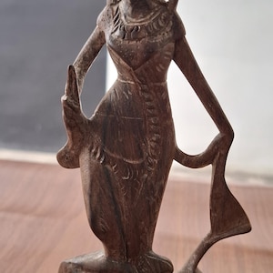 May include: A detailed wooden sculpture of a Balinese dancer in a traditional pose. The figure is carved from dark wood, showcasing intricate details of the costume and headdress. The sculpture is on a wooden surface.