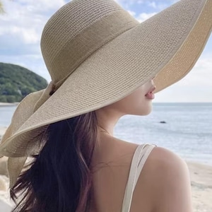 May include: A wide-brimmed sun hat in a natural beige color, featuring a matching band and a delicate bow. The hat's design offers ample shade, perfect for sunny days. The hat is made of woven material.