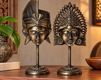 May include: Two bronze-toned statues featuring human faces on thin metal stands with circular bases. The left statue wears a turban-style headpiece, while the right statue features an ornate, crown-like headdress with intricate filigree detailing.