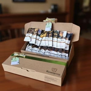 Può includere: Una confezione regalo in carta kraft, legata con un nastro verde e un piccolo cartellino. All'interno, sono disposti vari pacchetti di caffè. La scatola è aperta, rivelando il contenuto, ed è presentata su una superficie di legno.