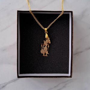 Puede incluir: Un collar con colgante dorado con una figura estilizada y detallada. El colgante cuelga de una delicada cadena y se presenta en una caja negra. El collar probablemente está hecho de metal dorado.