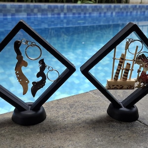 May include: Two black framed display cases with Indonesian keychains. One case features gold and black keychains, the other has a miniature puppet show scene. The cases are on a stone surface with a pool in the background.
