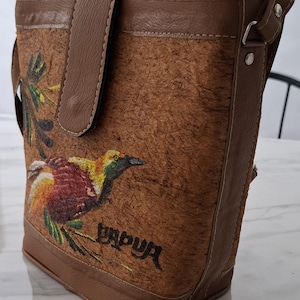 Könnte beinhalten: Braune Umhängetasche mit Klappenverschluss und langem Riemen. Die Tasche hat ein gemaltes Vogeldesign mit dem Wort "PAPUA" in Schwarz. Die Tasche besteht aus einem strukturierten Material mit braunen Lederbesätzen.