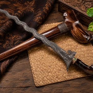 Op de afbeelding: Een dolk met een golvend lemmet, een donkere, getextureerde metalen afwerking en een gebogen handvat van gepolijst donker hout. Het handvat heeft een kleine decoratieve band met groene accenten. Inclusief een bijpassende schede van donker hout.