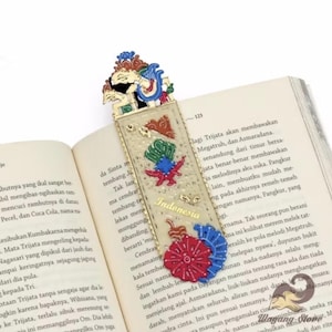 May include: A decorative bookmark featuring Indonesian cultural elements. The bookmark has a gold-colored top with a detailed figure, and a clear body adorned with colorful fabric designs and the word "Indonesia". It is placed in an open book.