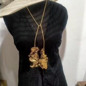 Può includere: Una collana color oro con un ciondolo raffigurante due figure. La collana è appoggiata su un top di pizzo nero. Un cappello di paglia a tesa larga è appoggiato sopra. La collana è un pezzo forte.