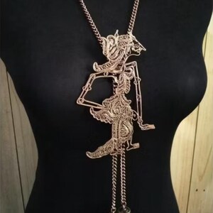 May include: A copper-toned pendant necklace with a detailed, filigree puppet design. The pendant hangs from a chain and has two decorative, bell-shaped charms at the bottom. The necklace is displayed on a black mannequin.
