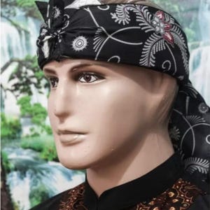 May include: A black and white patterned head wrap with a decorative knot, worn by a mannequin. The head wrap features a floral and geometric design. The mannequin is wearing a black shirt with a brown and gold patterned design.